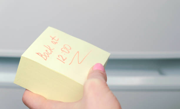 istock Yellow sticker paper sheet with words back at 12 in woman's hand. 987001206