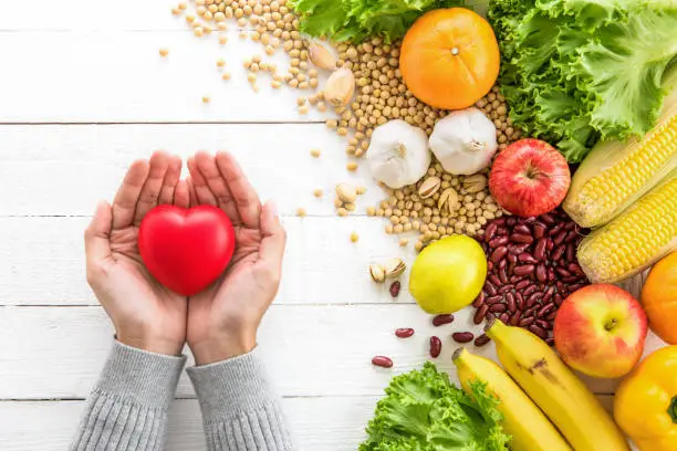 Woman hands holding red heart shape ball with various kinds of colorful healthy medicinal fruits, vegetables and nuts aside on white wood table, top view