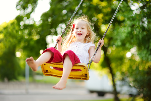 Cute little girl having fun on a playground outdoors on warm summer day. Cute kid swinging outdoors. Summer outdoor leisure for kids.