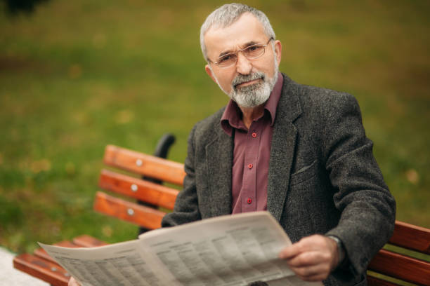 istock A nice grandfather with a beautiful beard in a gray jacket sits on a bench in the park and reads a newspaper. grandpa wear glasses 987262000