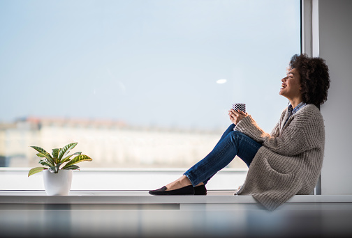 Young happy African American woman relaxing on a window sill during coffee time. Copy space.