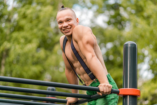 Cute young man with naked torso takes part in morning workout at outdoors gym in a public park. The young man doing bodyweight exercise. He is smiling looking at the camera. Shooting in sunny early morning at summer