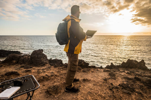 nomade digital voyageur homme au travail à la recherche d’un incroyable coucher de soleil doré sur l’océan. appareil photo et ordinateur portable et belle vie à des activités de loisirs en plein air. bureau de travail à l’abri - peuple nomade photos et images de collection