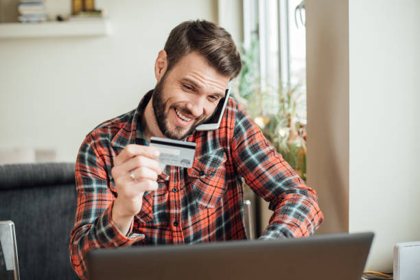 istock Smiling man using mobile phone while looking at credit card in his hand 987572646