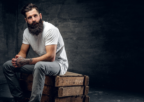 Bearded brutal male with tattoo on arm dressed in a white t shirt sits on a wooden box.