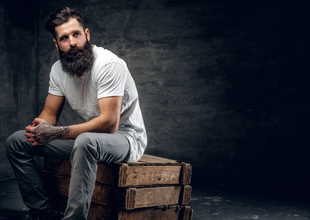 bearded male with tattoo on arm dressed in a white t shirt sits - arm tattoo imagens e fotografias de stock