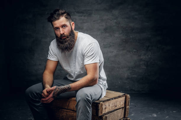 istock Bearded male with tattoo on arm dressed in a white t shirt sits 987615580