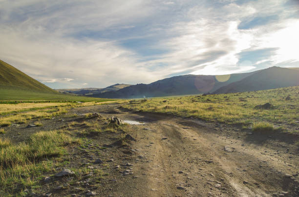 altai mongolo. strada sterrata e valle di montagna tra le travi del sole della sera. camino fumante dei nomadi yurta tradizionali. natura e viaggi. mongolia - dirt road foto e immagini stock