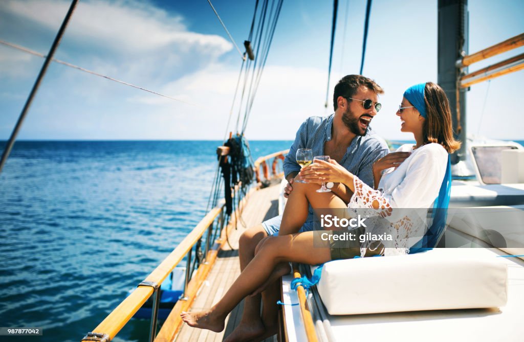 Couple on a sailboat. Closeup side view of a young couple relaxing on a sailboat cruise while sailing close to the coast. They are having some wine and laughing. Couple - Relationship Stock Photo
