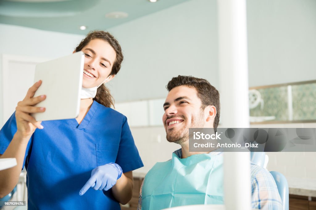 Smiling Dentist And Patient Looking At Digital Tablet In Clinic Smiling dentist and male patient looking at digital tablet during treatment in dental clinic Dentist Stock Photo