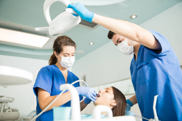 Dentist With Male Colleague Treating Patient At Clinic Dentist treating patient with dental tool while male colleague adjusting light at clinic dental light stock pictures, royalty-free photos & images