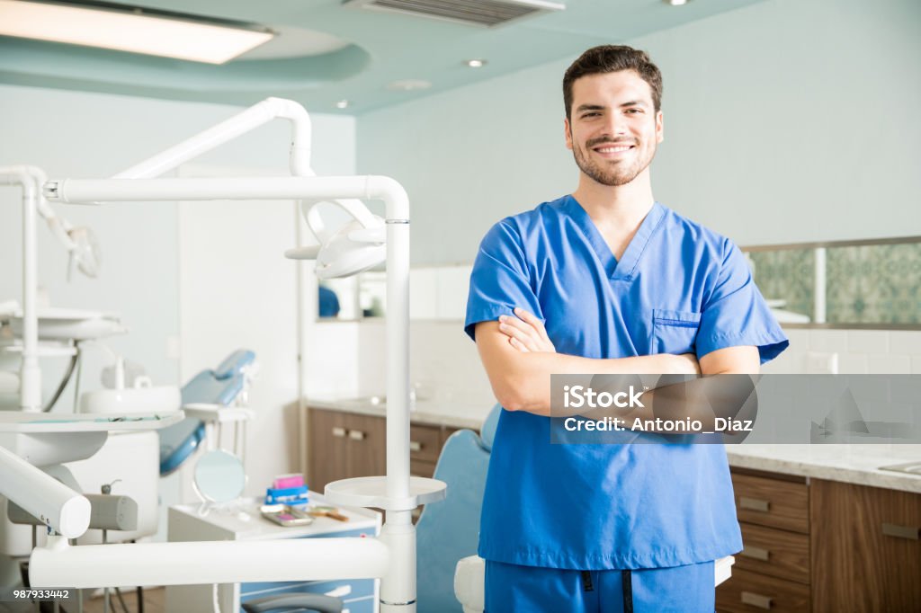 Portrait Of Confident Dentist Standing With Arms Crossed In Clinic Portrait of smiling confident mid adult dentist standing with arms crossed in dental clinic Dentist Stock Photo