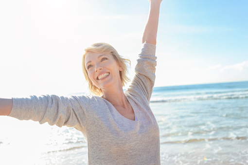 Mature woman on the beach with arms up. She looks very happy and relaxed. She is smiling and joyful with the ocean behind her. Bright sunny summer day