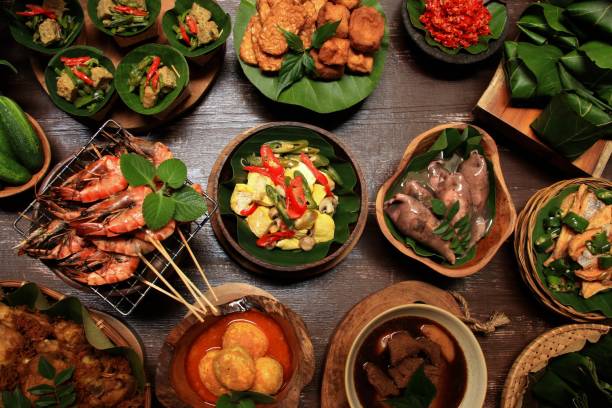 Nasi Jamblang, the popular, traditional rice meal from Cirebon, West Java. Parcels of steamed rice wrapped in fragrant teak leaf served with plenty, optional side dishes, such as Braised Squids, Soy Beef Stew, Curry Egg, Fried Chicken, Fried Tempeh and Tofu, Crispy Salted Fish, Yellow Tofu, Prawn Satay, Sautéed Velvet Leaf, and Mushrooms in Banana Leaf Pouch. A typical condiment for this meal, Sambal Iris or Sliced Chili Salsa, is also served along. The dishes are plated in various kind of crockery and arranged on a slightly rustic wooden table.