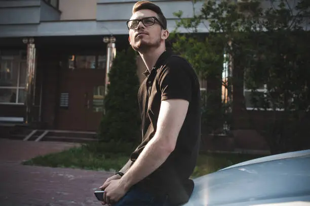 Photo of Young man leaning on his car, using a smartphone, dressed casually.
