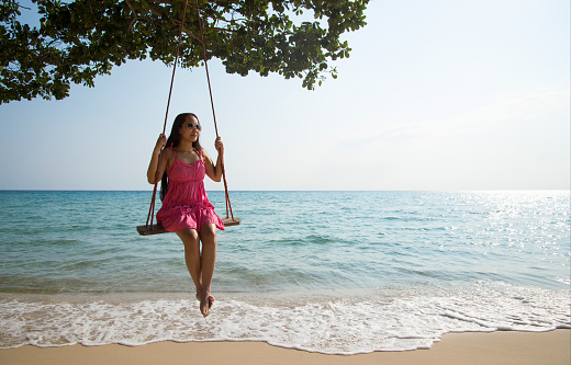 A beautiful young Asian woman relaxes sitting on a rope swing on an idyllic tropical beach.