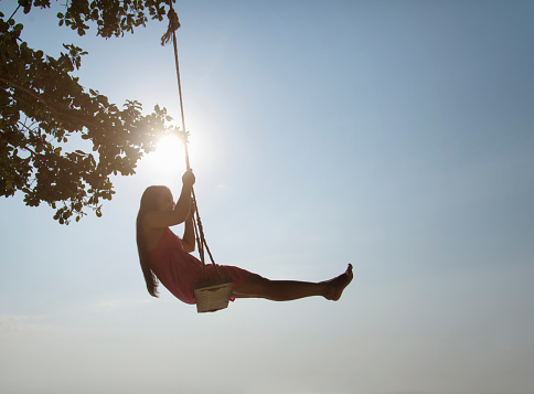 A young Asian woman plays on a rope swing silhouetted against a sunburst through the tree leaves.