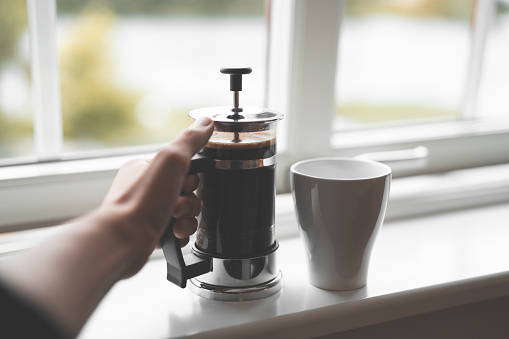 Personal perspective of a man with a french style coffee press filled with hot coffee on a white window sill inside a house in Sweden. Close-up shot with selective focus.