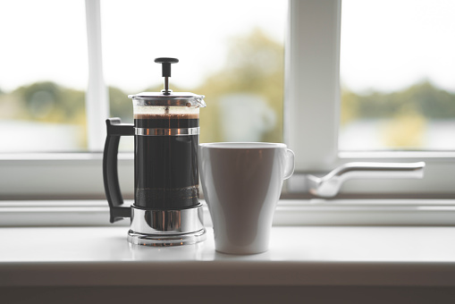 French style coffee press filled with hot coffee on a white window sill inside a house in Sweden. Close-up shot with selective focus.