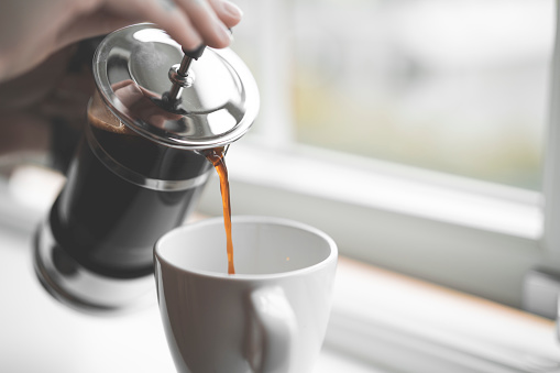 French style coffee press filled with hot coffee on a white window sill inside a house in Sweden. Close-up shot with selective focus of a person with tattoos pouring coffee into a cup.