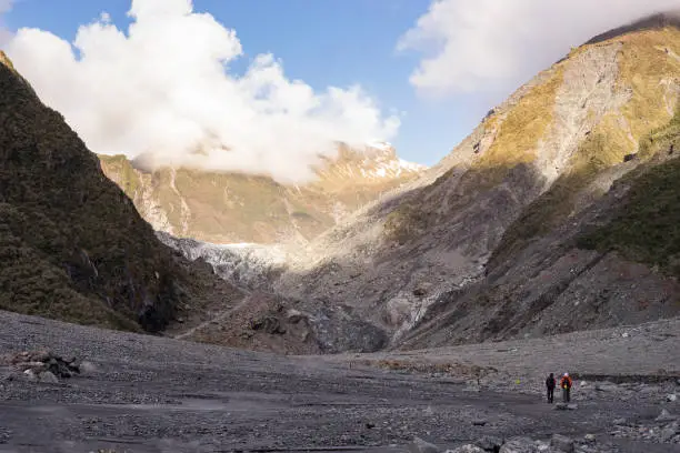 Photo of landscape of huge mountains with tow small people walk on the way to fox glacier in New Zealand.