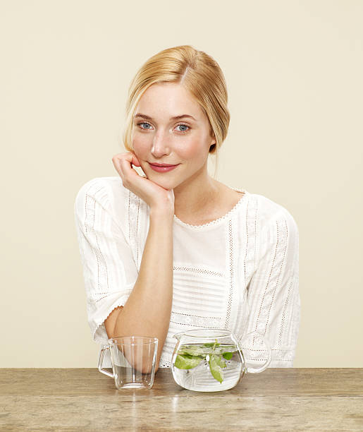 female smiling with fresh brewing tea female, blonde hair, blue eyes, smile, white dress, fresh tea, glass tea pot, glass cup, biscuit background, yellow background hand on chin stock pictures, royalty-free photos & images