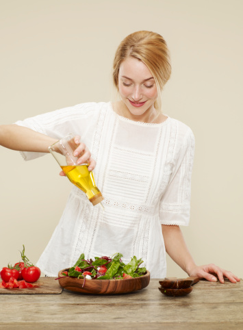 female, blonde hair, blue eyes, smile, white dress,fresh salad, salad bowl, mixing tongs mixed leaf, sliced tomoatoe, olive oil, oil drizzler, cutting board, biscuit background, yellow background