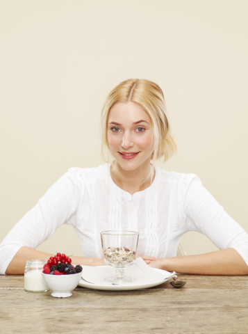 female, blonde hair, blue eyes, smile, white top,cherries, blueberries, blackberries, raspberries, bowl of fruit, fruit and nut cereal, cereal, plate, spoon, jar of yoghurt, biscuit background, yellow background