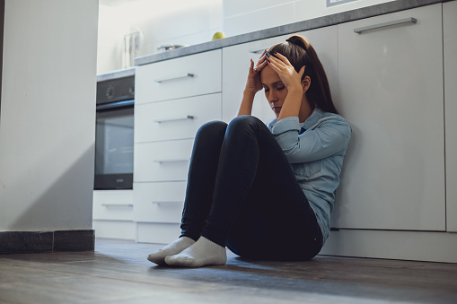 Depressed woman sitting on a kitchen floor and holding her head
