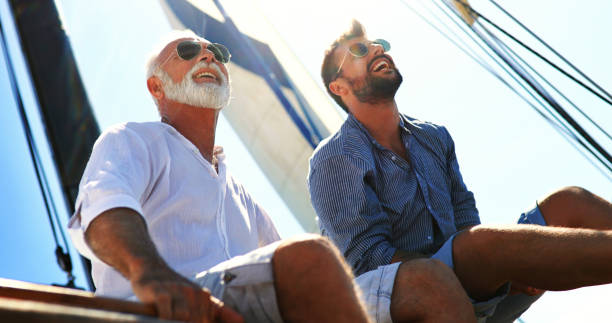 Father and son sailing. Closeup low angle view of mid 60's man and his son enjoying summer holiday on a sailboat. They are sitting on the deck, looking at the distance, talking and laughing. old rich white guy stock pictures, royalty-free photos & images
