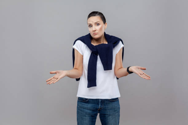 i don't know. handsome casual styled woman with blue jeans and sweater on her shoulders, smart watch on arm, looking at camera and confused. - blank expression head and shoulders horizontal studio shot imagens e fotografias de stock