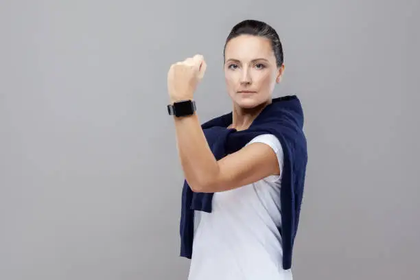 Photo of Portrait of dominating brunette woman in casual clothes with jeans and sweater on her shoulders looking serious at camera showing fist and smart watch.