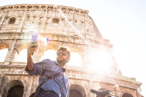 Photo of Young hipster man with bike at colosseum taking selfies pictures with smartphone in Rome city centre on sunny day
