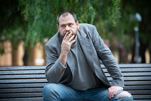 Adult Man sitting on a bench at public park smoke a cigarette
