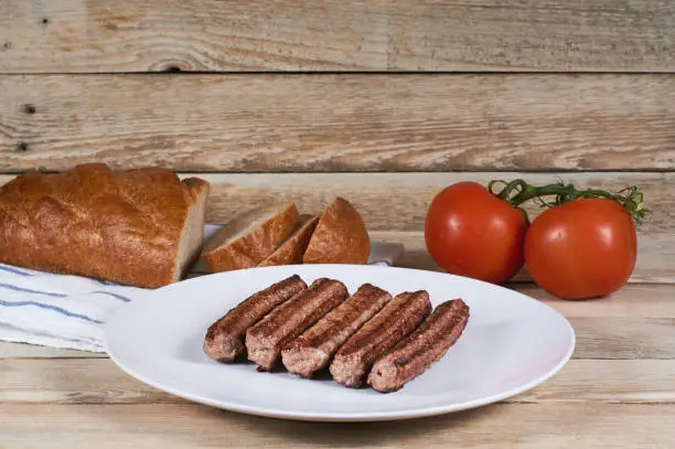 Serbian cevapi, cevapcici, Balkan minced meat kebab on a white plate with bread and tomatoes on a napkin and a wooden background