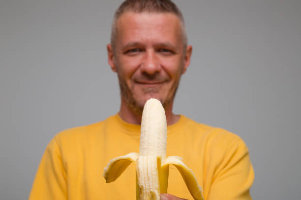 ritratto di uomo adulto che mangia tenendo la banana di fronte a lui al chiuso - men banana holding cut out foto e immagini stock