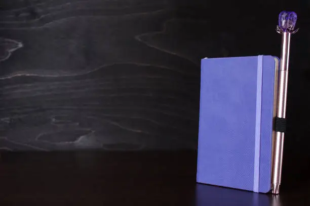 Photo of Purple notepad with an attached pen on the table on a black woodenl background