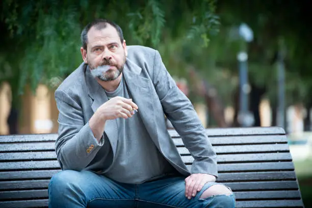 Photo of Adult Man sitting on a bench at public park smoke a cigarette