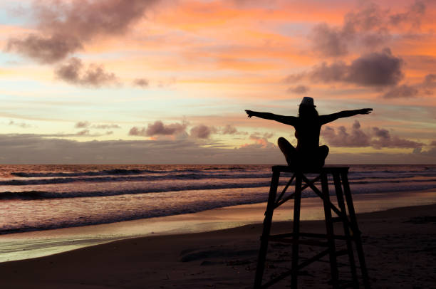 una mujer de silueta sentado en una torre de salvavidas con los brazos abiertos disfrutando del amanecer en una playa y el sol pintando el cielo con colores anaranjados. - lifeguard orange nature beach fotografías e imágenes de stock