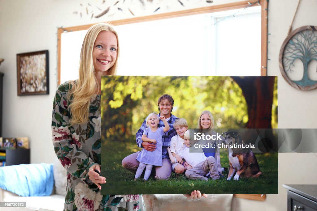 Happy Young Woman Holding Canvas Print of Family Portrait A happy young blonde woman is holding a large wall canvas portrait of her family with young children and a pet dog. Artist's Canvas Stock Photo