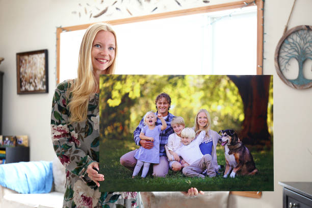 Happy Young Woman Holding Canvas Print of Family Portrait A happy young blonde woman is holding a large wall canvas portrait of her family with young children and a pet dog. animal related occupation photos stock pictures, royalty-free photos & images