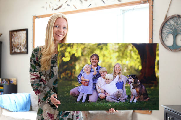 Happy Young Woman Holding Canvas Print of Family Portrait A happy young blonde woman is holding a large wall canvas portrait of her family with young children and a pet dog. printmaking technique stock pictures, royalty-free photos & images