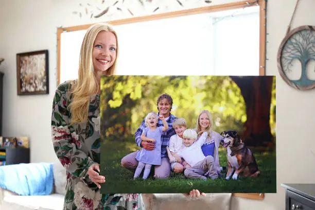 A happy young blonde woman is holding a large wall canvas portrait of her family with young children and a pet dog.