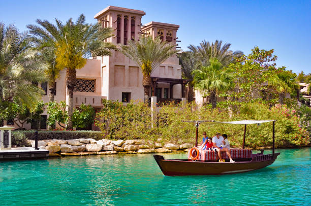 a man, a woman and children ride an old wooden boat along the canal at madinat past the buildings of arab culture - madinat jumeirah hotel imagens e fotografias de stock
