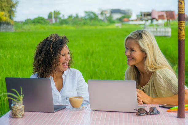 istock two happy female friends working outdoors at beautiful internet cafe with laptop computer , a caucasian woman and an afro mixed ethnicity girl enjoying together as digital nomad girlfriends 989002838