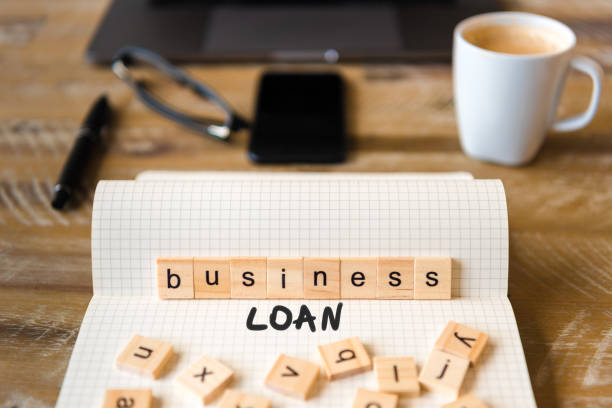 istock Closeup on notebook over vintage desk background, front focus on wooden blocks with letters making Business Loan text 989125130
