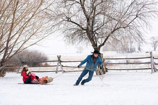Caucasian cowboy wearing a denim jacket, cowboy boots and black hat pulls his family on a wooden tobaggon by a rope across the snow in front of a rustic fence with bare trees on a snowy subzero cold windy day on a horse ranch, Livingston, Montana, USA