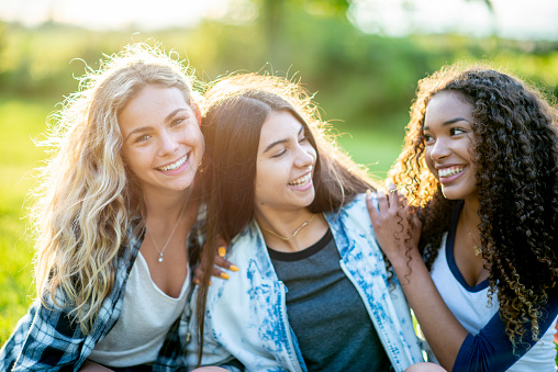 Three teenage girls of different ethnicity are outdoors. They are embracing and smiling on this sunny day.