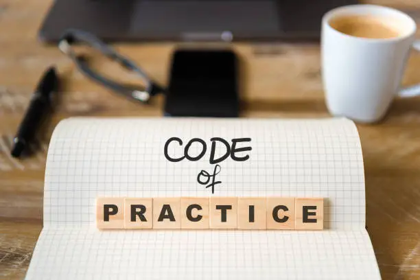 Closeup on notebook over wood table background, focus on wooden blocks with letters making Code of Practice text. Concept image. Laptop, glasses, pen and mobile phone in defocused background