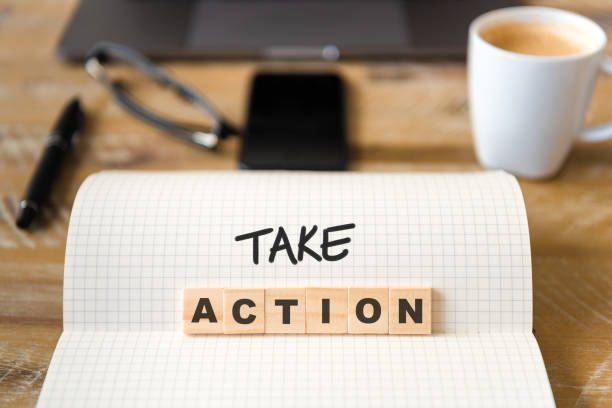 istock Closeup on notebook over wood table background, focus on wooden blocks with letters making Take Action text 989180094